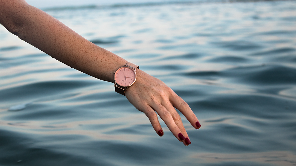 A close up shot of someone's arm being held out against a background of water. On the wrist is a pink dive watch.