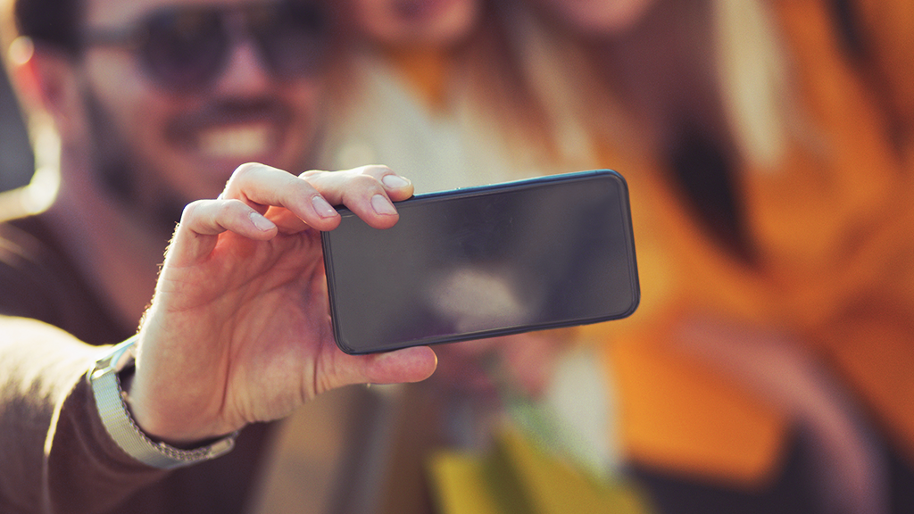 A close up shot of a hand holding a smart phone as if to take a selfie of the blurred man in the background.
