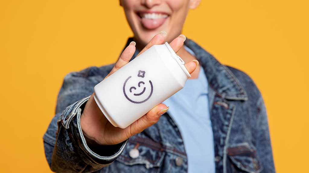 A smiling woman thrusting a can of soda at the viewer. The can is white with a logo of a smiling face. 