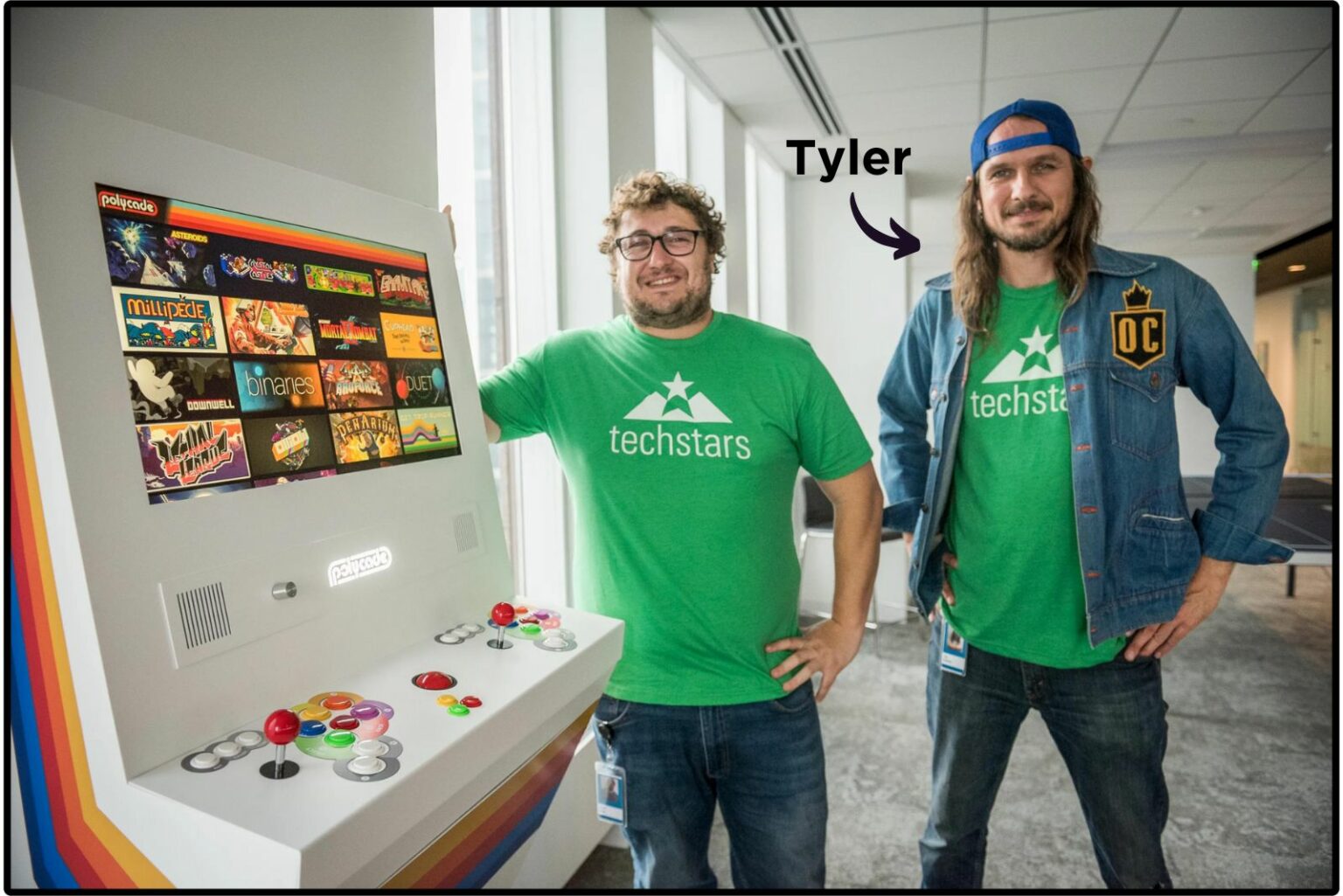 Two bearded men in green "techstars" tee-shirts standing next to a poster covered in cabinet art from vintage arcade games. An arrow from the name "Tyler" is pointing to the taller one wearing a hat and jean jacket. 